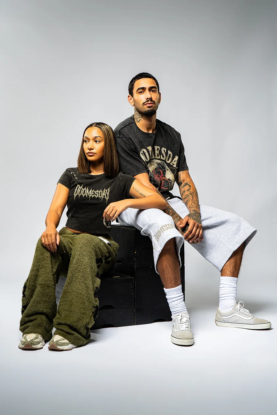 Two seated models in DOOMESDAY tees, olive textured trousers, and grey long shorts, showing complete Survive.The.Day looks.
