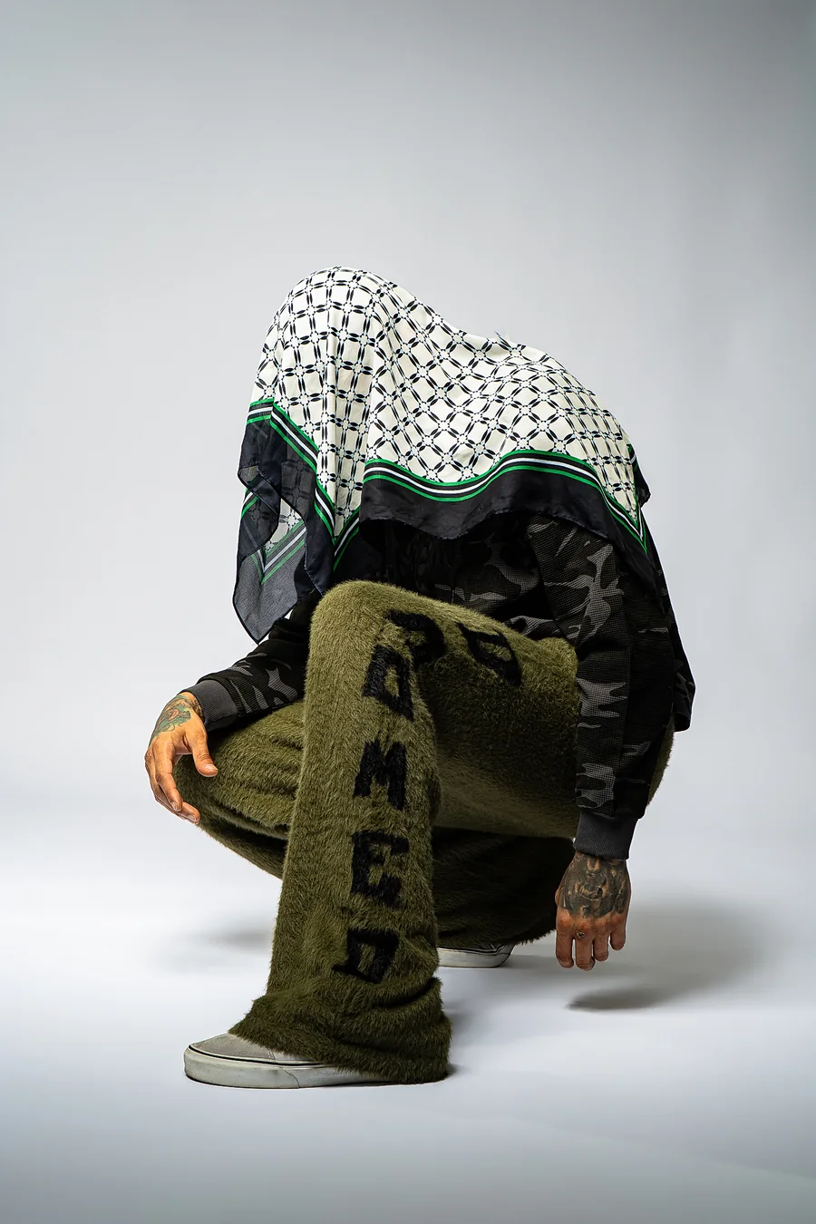 Crouching model in olive textured trousers and a camouflage top with a black, white, and green scarf draped over their head.