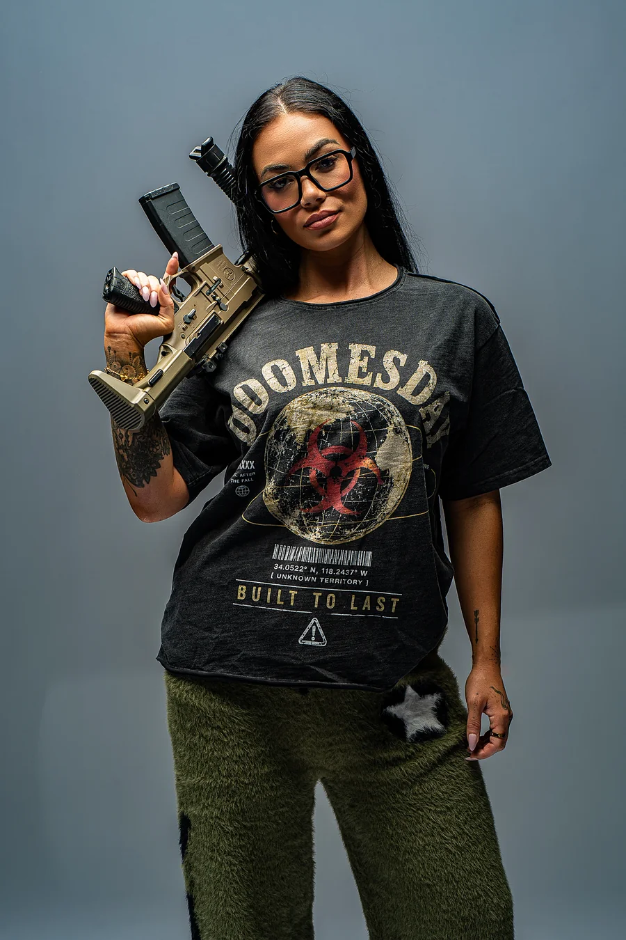 Model in an oversized black DOOMESDAY graphic tee and olive trousers, holding a firearm over one shoulder.